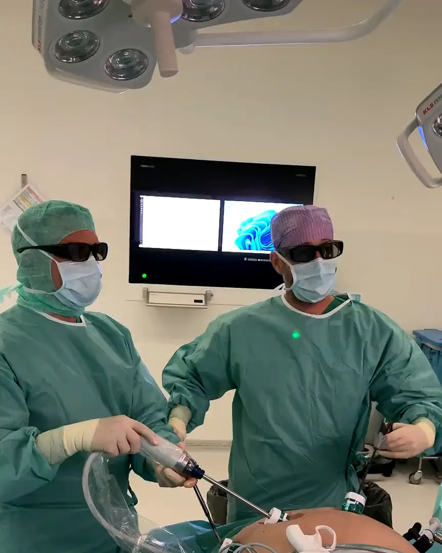 Zwei Chirurgen mit 3D-Brillen operieren in Schlüssellochtechnik, dahinter der Monitor